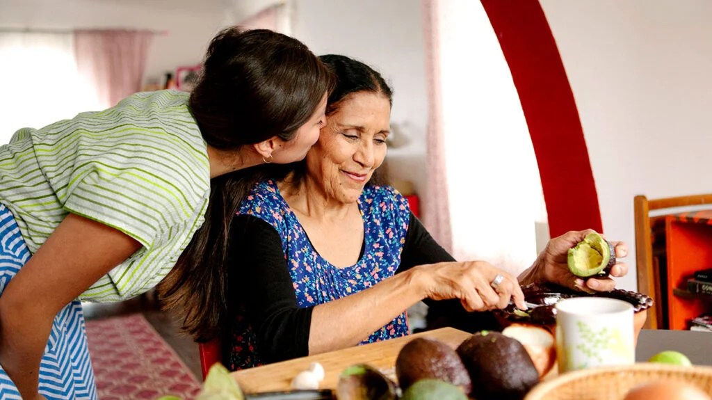 A woman kisses the cheek of an older woman who's cutting avocados